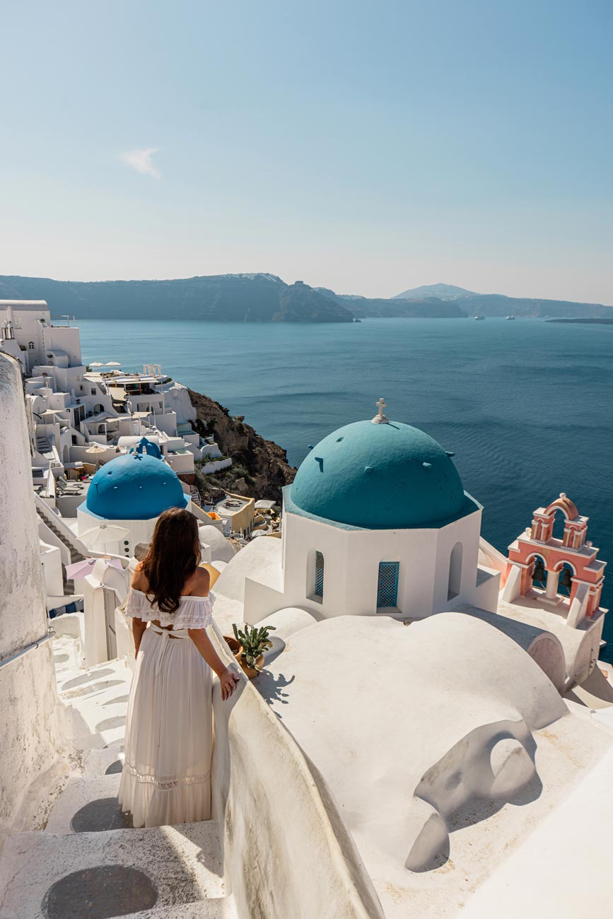 A woman in a white dress standing on steps overlooking the iconic blue-domed churches of Oia, Santorini, with whitewashed buildings and the deep blue Aegean Sea stretching into the horizon.