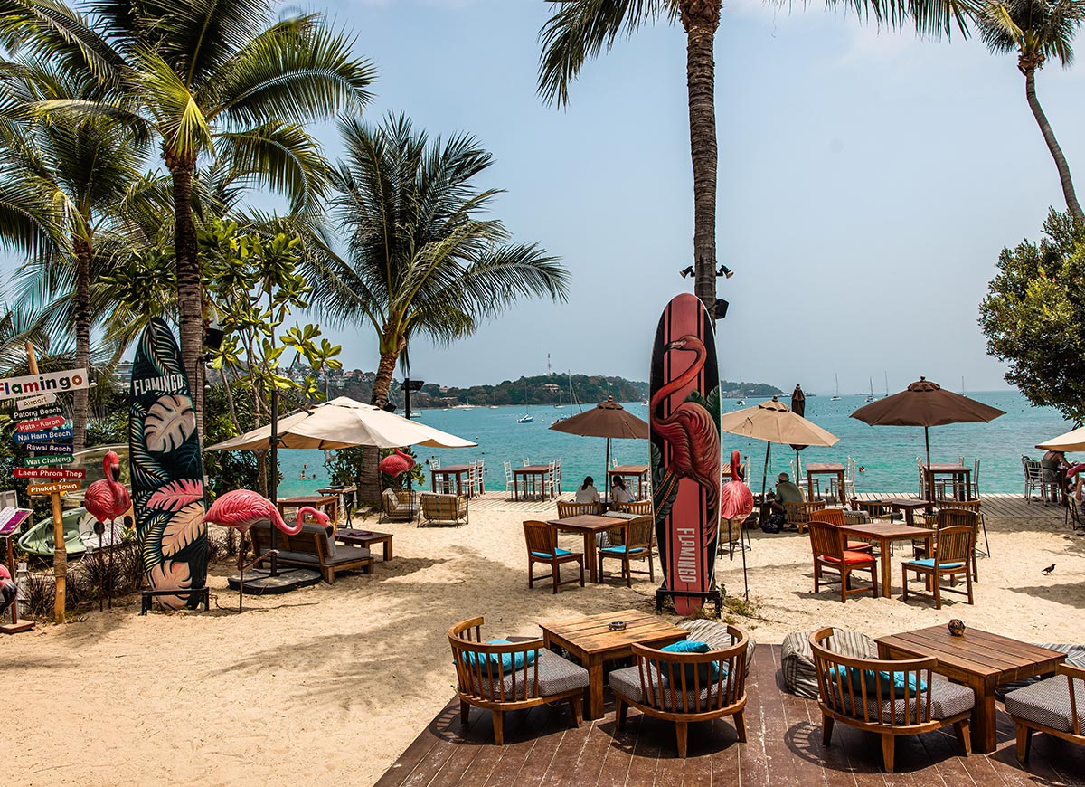 Beachfront setting with wooden chairs and tables arranged on sand, surrounded by palm trees, flamingo-themed decorations, and surfboards, overlooking a calm turquoise sea with boats in the distance.