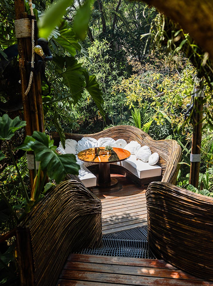 This is an image of an outdoor seating area surrounded by lush greenery. The setup features a cozy circular arrangement of white-cushioned seats around a polished wooden table, framed by intricately woven wooden railings and shaded by tropical plants. The inviting atmosphere highlights a blend of natural and crafted aesthetics, ideal for a relaxing retreat.