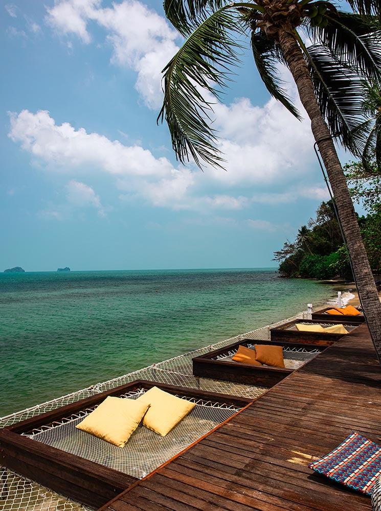 A wooden deck along the shoreline features built-in net hammocks with plush yellow pillows, offering a relaxing spot above the turquoise waters. A swaying palm tree frames the view of the ocean and distant islands under a bright blue sky.