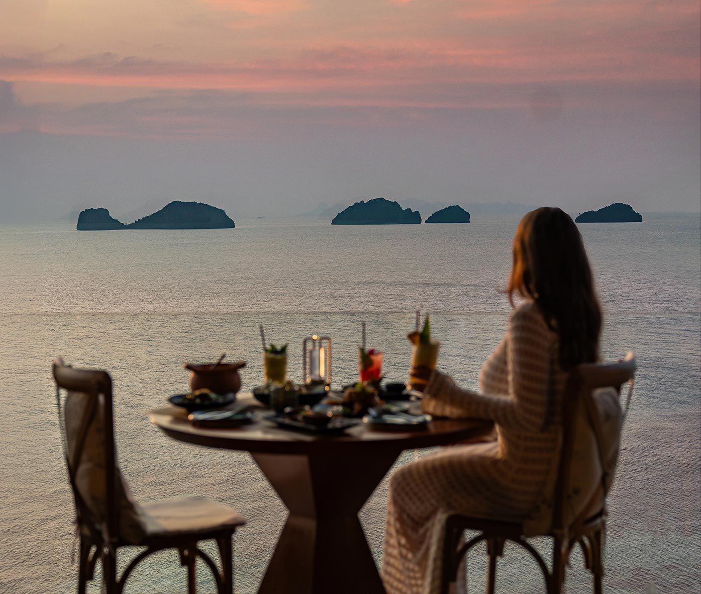 A woman in an elegant crochet dress enjoys a sunset dinner at a seaside table, gazing at the ocean and distant islands. The intimate setting features gourmet dishes, tropical cocktails, and a softly lit lantern, creating a serene and romantic dining experience.