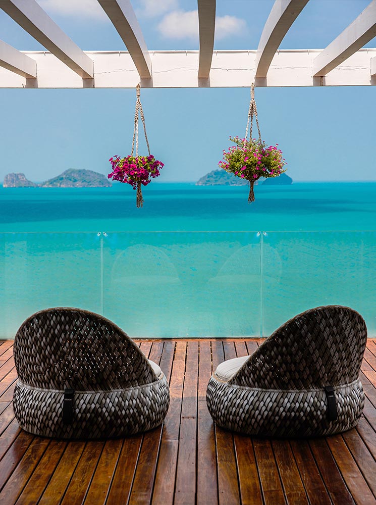 Two woven lounge chairs sit on a wooden deck overlooking the turquoise ocean, framed by a white pergola with hanging flower baskets. A clear glass barrier enhances the uninterrupted view of the distant islands under a bright blue sky.