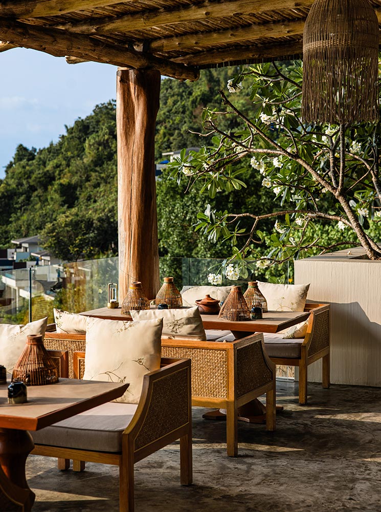 An outdoor restaurant with rustic wooden furniture and woven rattan details is set under a thatched roof, surrounded by lush greenery. Earth-toned cushions, decorative lanterns, and blooming frangipani trees create a serene and inviting dining atmosphere.