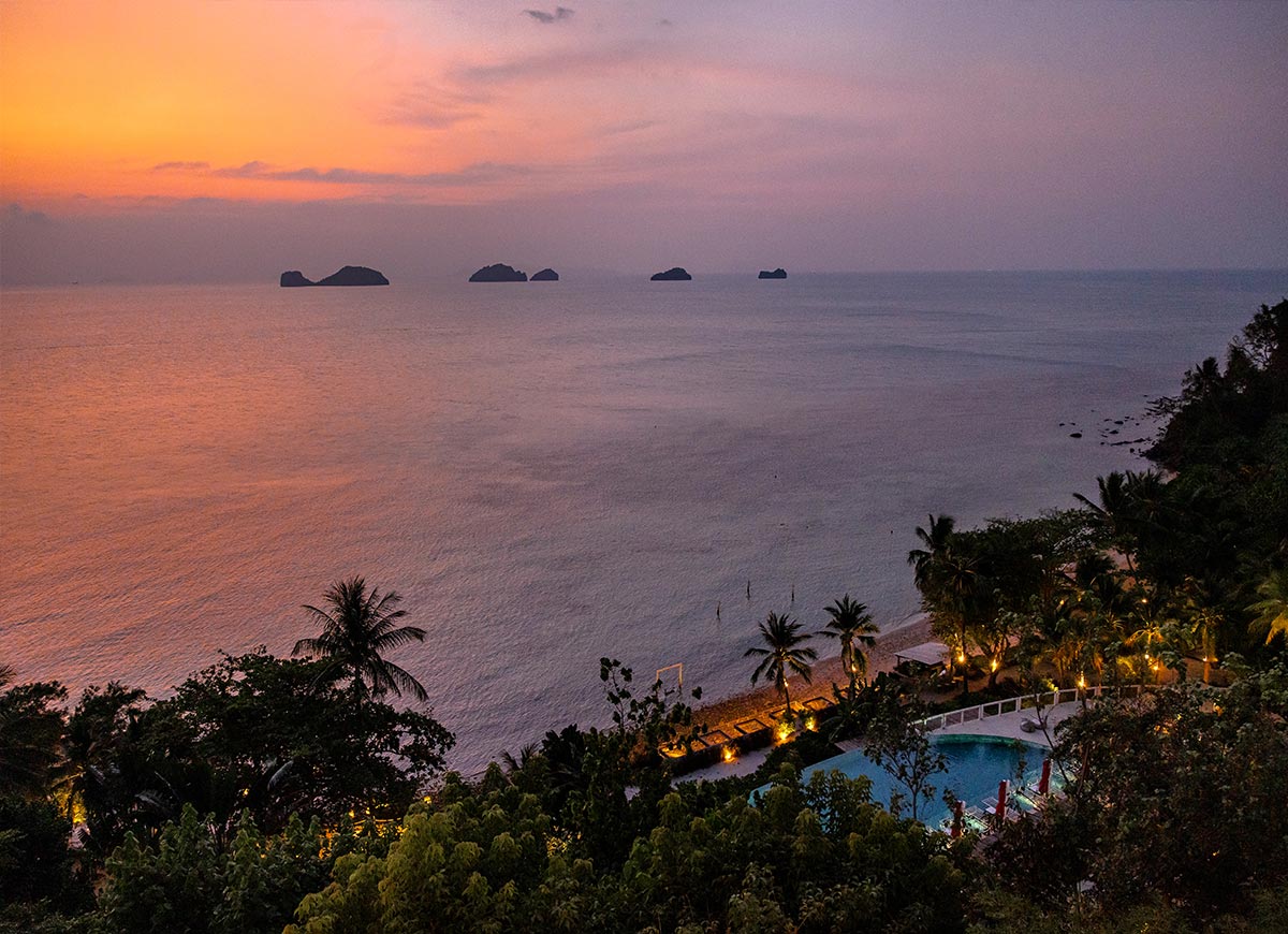 A breathtaking sunset casts warm hues of orange and pink over the calm ocean, with silhouettes of small islands in the distance. Below, a secluded beach and illuminated resort pool are nestled among lush tropical greenery, creating a serene and picturesque evening view.