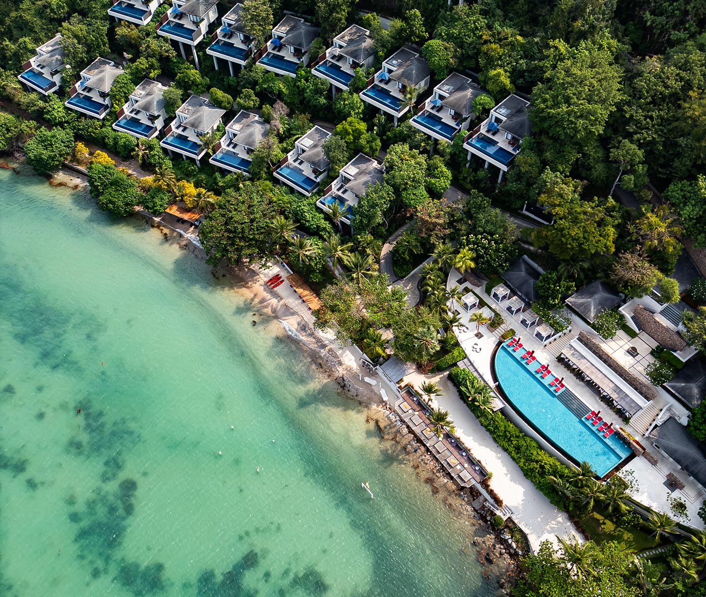 An aerial view of a luxurious beachfront resort showcases private villas with infinity pools nestled among lush greenery. The turquoise waters of the ocean meet a pristine sandy beach, with an expansive pool and red lounge chairs adding to the tropical paradise setting.