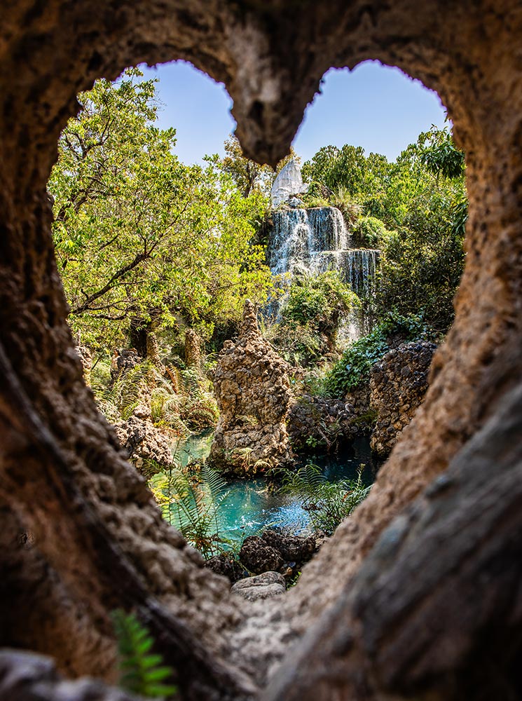 A cascading waterfall flows into a turquoise pool, framed by a heart-shaped opening in the rocky foreground. Lush green foliage surrounds the serene landscape, with jagged rock formations adding a mystical touch. The natural framing creates a dreamy, picturesque view of this hidden oasis.