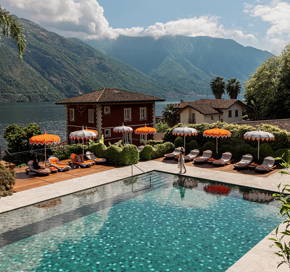 A luxurious poolside scene at Grand Hotel Tremezzo in Lake Como, Italy, featuring vibrant orange and white umbrellas, sun loungers, and a sparkling turquoise pool. A woman in a white dress walks along the deck, with the stunning backdrop of mountains, lush greenery, and the serene lake. The hotel's signature red villa adds a touch of charm to the picturesque setting.