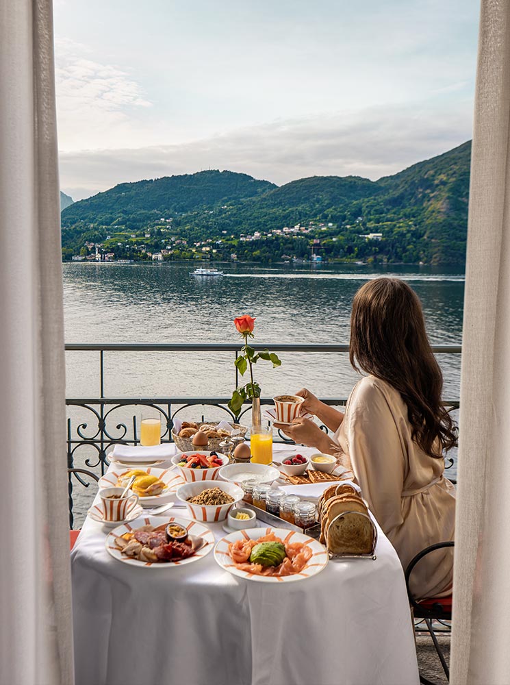 A woman in a silky robe enjoys a lavish breakfast spread on a balcony overlooking the serene waters of Lake Como. The table is adorned with fresh fruits, pastries, smoked salmon, and orange juice, with a single red rose as a centerpiece. Lush green hills and charming lakeside villages create a breathtaking backdrop for this idyllic morning scene.