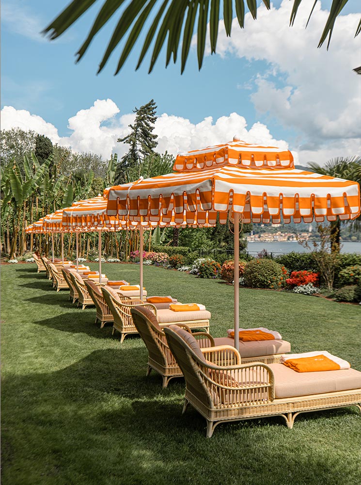 A row of elegant wicker sun loungers with orange cushions sits under matching striped umbrellas on a perfectly manicured lawn at Grand Hotel Tremezzo. The lush greenery, vibrant flowers, and palm trees frame a picturesque view of Lake Como in the background. The luxurious setting exudes relaxation and timeless Italian charm.