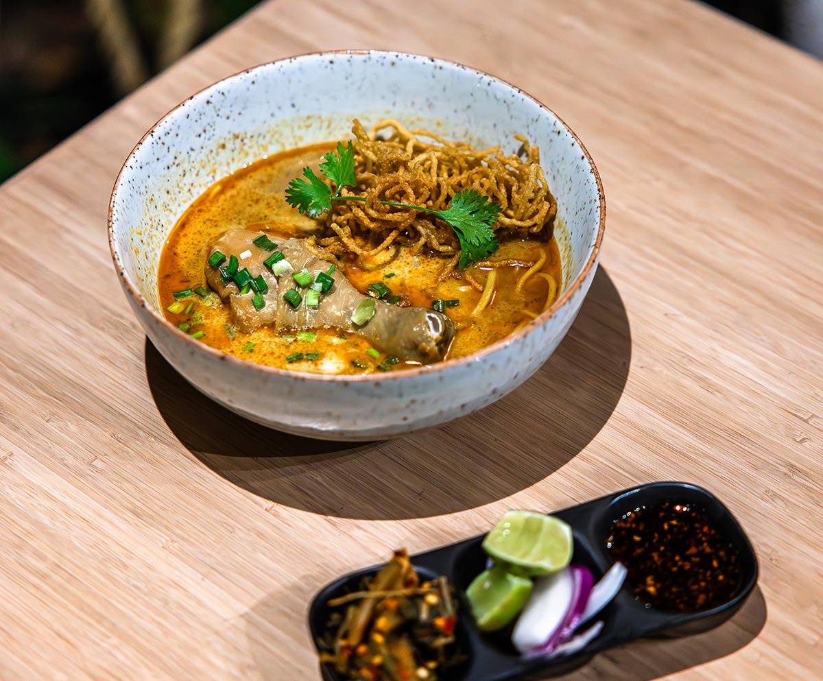 A bowl of Khao Soi, a Northern Thai coconut curry noodle soup, served in a speckled ceramic bowl. The dish features a golden broth with a chicken drumstick, crispy fried noodles, fresh cilantro, and chopped green onions. A black tray beside the bowl holds traditional condiments, including pickled mustard greens, lime wedges, sliced red onion, and a dark chili oil sauce. The meal is presented on a light wooden table, highlighting the vibrant colors and textures of the dish.


