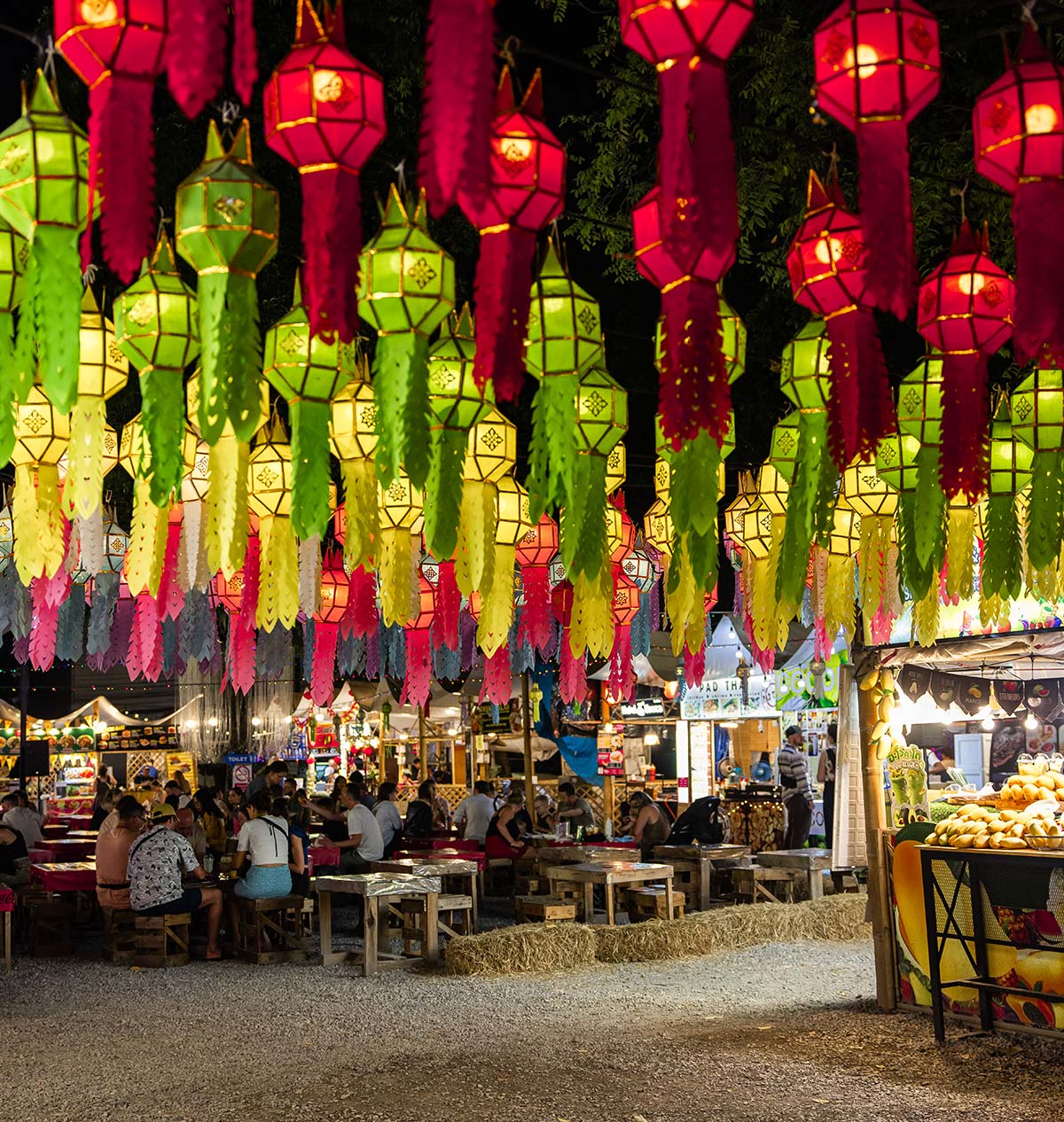 A vibrant night market scene is illuminated by colorful Lanna-style lanterns in red, green, and yellow, hanging in rows from above. Below, people sit at rustic wooden tables, enjoying food and conversation, while market stalls display fresh fruits and various goods. The lively atmosphere, warm lighting, and traditional decorations create an inviting and festive ambiance.






