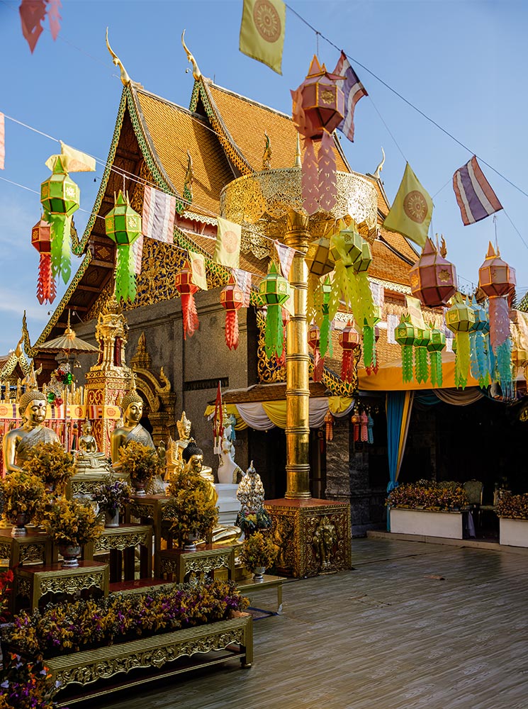 A beautifully adorned Buddhist temple courtyard featuring golden Buddha statues, vibrant flower arrangements, and colorful Lanna-style lanterns hanging overhead. The temple’s traditional Thai architecture, with its intricate gold detailing and sloping roofs, is illuminated by the warm sunlight. Flags flutter in the breeze, adding to the festive and spiritual ambiance of this sacred site.