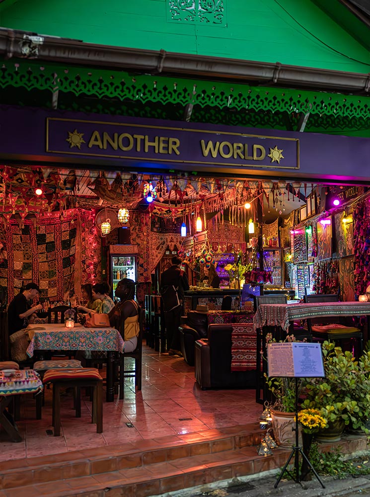 A vibrant and eclectic restaurant named "Another World" in Chiang Mai, Thailand, with an inviting atmosphere. The interior is adorned with colorful fabrics, hanging lanterns, and intricate decorations, creating a cozy and bohemian vibe. Diners enjoy their meals at patterned tables while warm lighting enhances the intimate and unique dining experience.