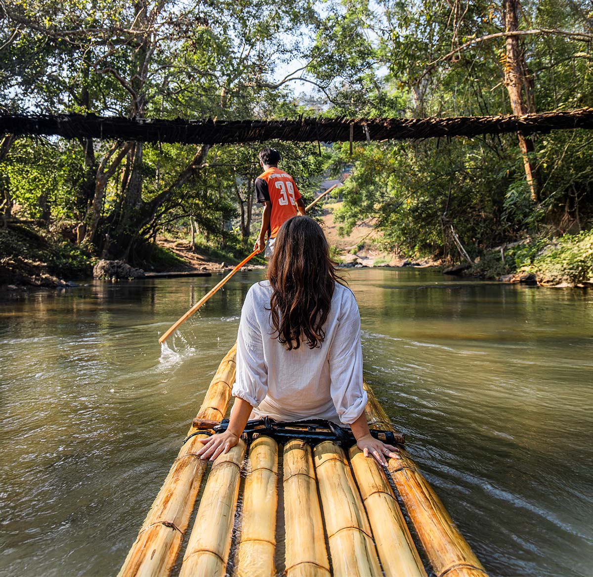 A woman in a white shirt sits on a traditional bamboo raft, gliding down a calm river surrounded by lush greenery. A local guide, wearing an orange jersey, stands at the front, using a long wooden pole to navigate the gentle waters. Sunlight filters through the trees, casting a warm glow over this peaceful and immersive nature experience in Chiang Mai, Thailand.
