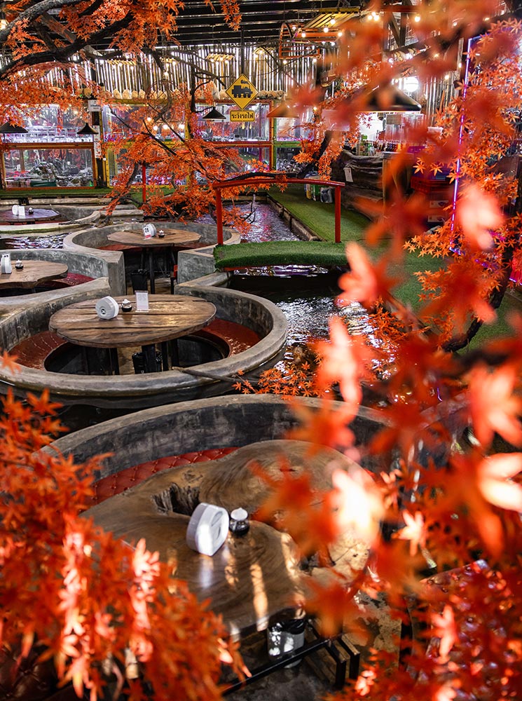 A cozy café with round, sunken seating areas surrounded by water, creating a unique dining experience. Vibrant orange maple leaves frame the scene, while a red bridge and koi ponds add to the tranquil ambiance. The warm lighting and rustic wooden tables enhance the inviting and immersive atmosphere of this koi café in Chiang Mai, Thailand.