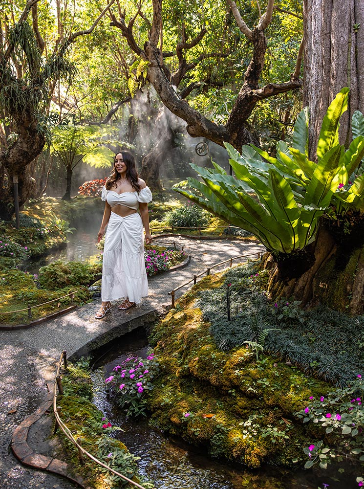 A woman in a flowing white dress walks along a curved stone path in a lush, misty garden. Towering trees, vibrant green ferns, and moss-covered rocks surround a gently flowing stream lined with delicate pink flowers. The soft sunlight filtering through the branches creates a magical, fairytale-like atmosphere in this serene setting.