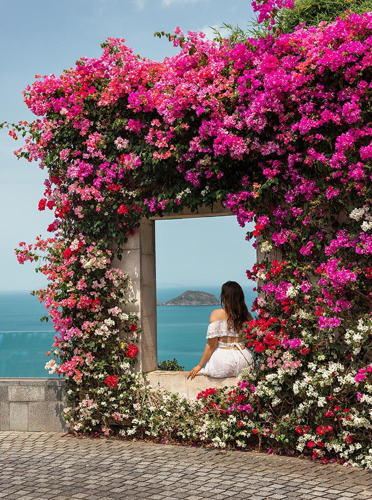 A woman in a white off-shoulder dress sits on the edge of a stone window framed by vibrant pink, red, and white bougainvillea flowers, gazing at the turquoise ocean and a distant island under a clear blue sky.