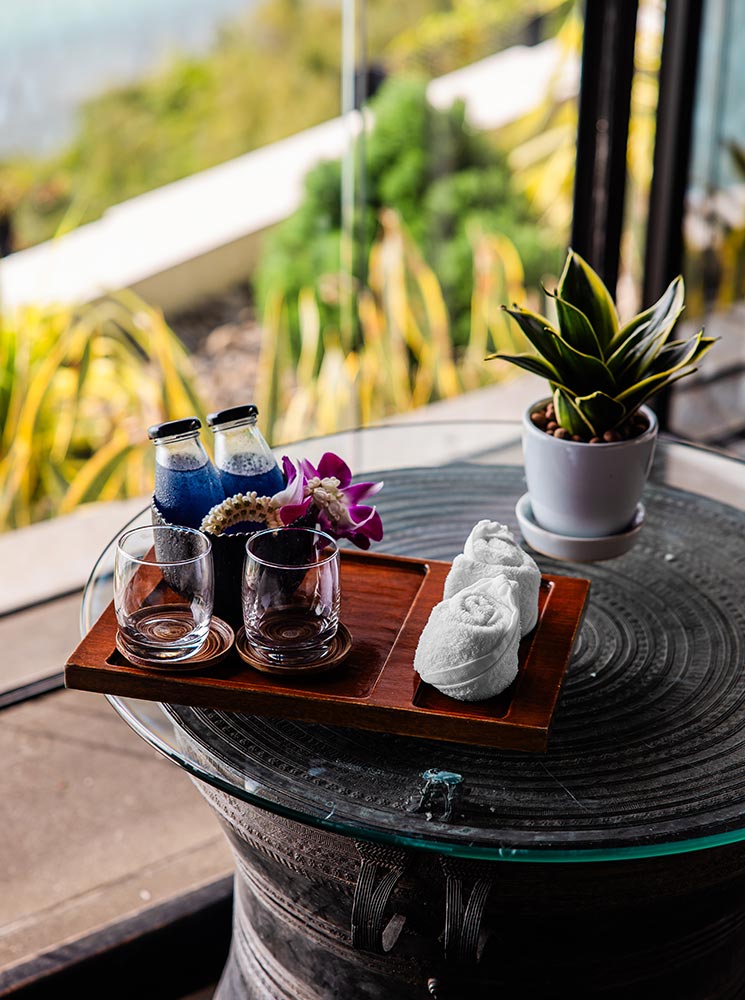 A wooden tray on a glass table holds two bottles of vibrant blue welcome drinks, two empty glasses, and neatly rolled white towels, accented with a purple orchid. A small potted plant sits beside the tray, with a blurred outdoor view in the background.