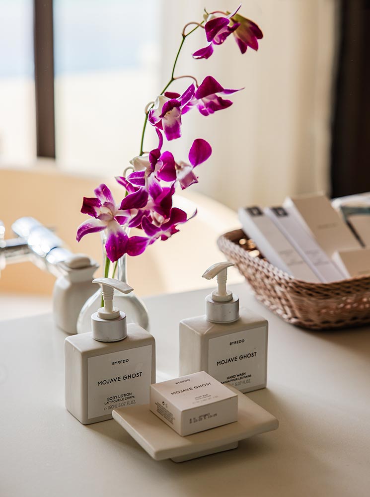 A close-up of Byredo Mojave Ghost bath products, including body lotion, hand wash, and soap, neatly arranged on a bathroom countertop. A vase with vibrant purple orchids and a wicker basket with amenities add a touch of elegance to the setup.