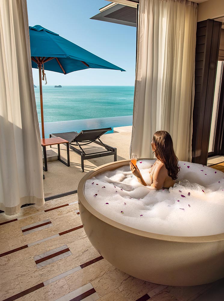 A woman relaxes in a luxurious round bathtub filled with bubbles and sprinkled with flower petals while sipping a cocktail. The open-air villa bathroom offers a breathtaking view of the turquoise ocean through sheer curtains. Outside, a sun lounger and a blue umbrella sit on the private terrace beside an infinity pool overlooking the sea.