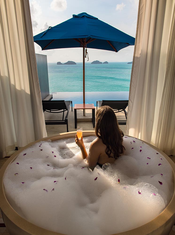 A woman relaxes in a bubble-filled bathtub sprinkled with flower petals, holding a glass of orange juice while gazing at the turquoise ocean. The open-air villa features flowing white curtains, an infinity pool, and a shaded lounge area with a blue umbrella overlooking the sea.