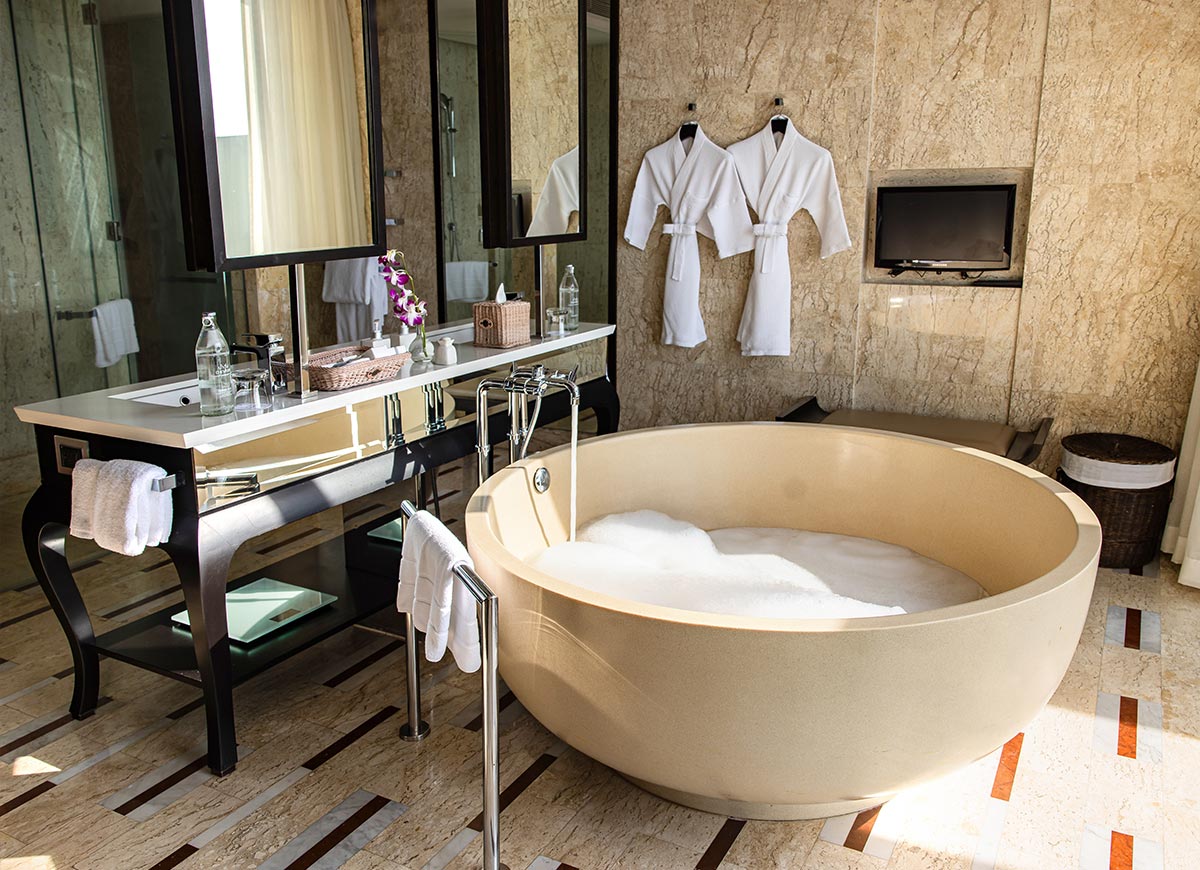 A luxurious bathroom features a large round soaking bathtub filled with bubbles, a sleek vanity with a mirror, and two plush white robes hanging on the marble wall. Sunlight streams in, highlighting the elegant design and warm tones of the space.