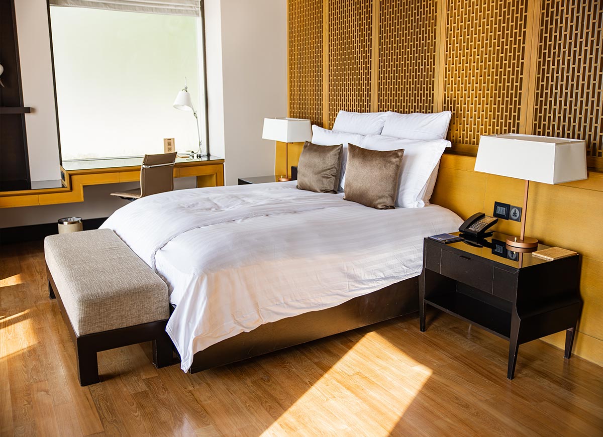 A luxurious hotel bedroom features a plush white bed with brown accent pillows, a wooden headboard with a geometric pattern, and modern bedside tables with lamps. A sleek desk with a chair sits by a large frosted window, adding to the room's elegant and contemporary design.