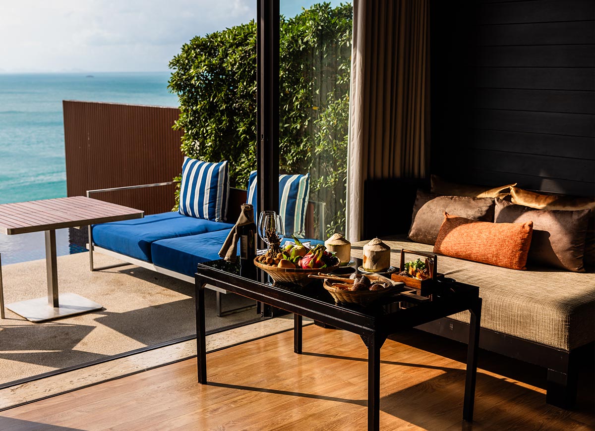 A luxurious villa living area features a table set with an assortment of tropical fruits, coconuts, and gourmet snacks, with a bottle of champagne and glasses. A plush daybed with decorative pillows sits beside a sliding glass door, leading to a private terrace with a blue cushioned lounge and an ocean view.