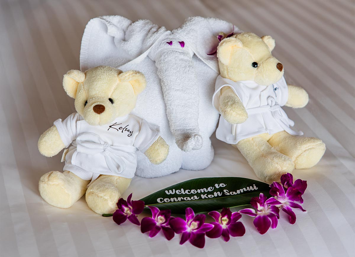 Two plush teddy bears dressed in miniature white robes sit beside a towel elephant on a neatly made bed. A green leaf with "Welcome to Conrad Koh Samui" written on it is decorated with vibrant purple orchids, creating a warm and personalized welcome arrangement.