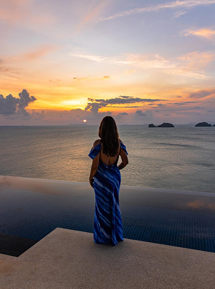 A woman in a flowing blue dress stands at the edge of an infinity pool, gazing at the sunset over the ocean. The sky is painted in shades of orange, pink, and purple, reflecting on the calm water. Small islands dot the horizon, adding to the serene and picturesque view.