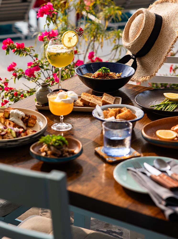 A beautifully set outdoor dining table features an assortment of gourmet dishes, including pasta, grilled seafood, fresh vegetables, and bread, paired with refreshing citrus cocktails. A straw sun hat rests on the chair, with vibrant pink flowers and a poolside view creating a relaxed tropical ambiance.