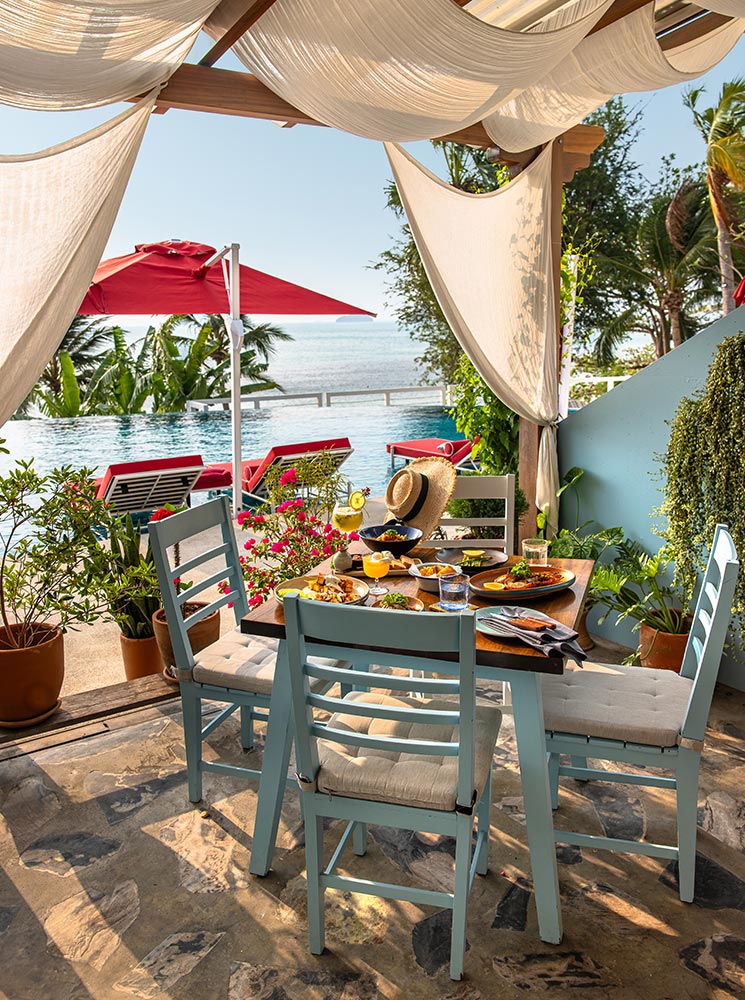 A picturesque outdoor dining setup features a wooden table with light blue cushioned chairs under a flowing white canopy. A delicious meal with fresh tropical dishes is served, overlooking an infinity pool with red lounge chairs and a stunning ocean view.