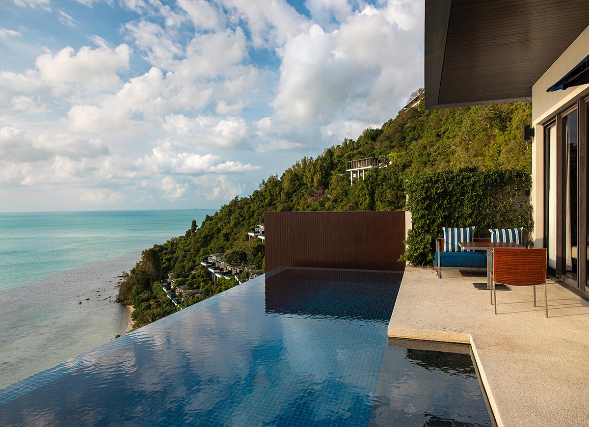 A private infinity pool overlooks the turquoise ocean and lush hillside villas, creating a serene retreat. The villa’s outdoor terrace features a cozy seating area with striped cushions, surrounded by greenery and modern architecture.