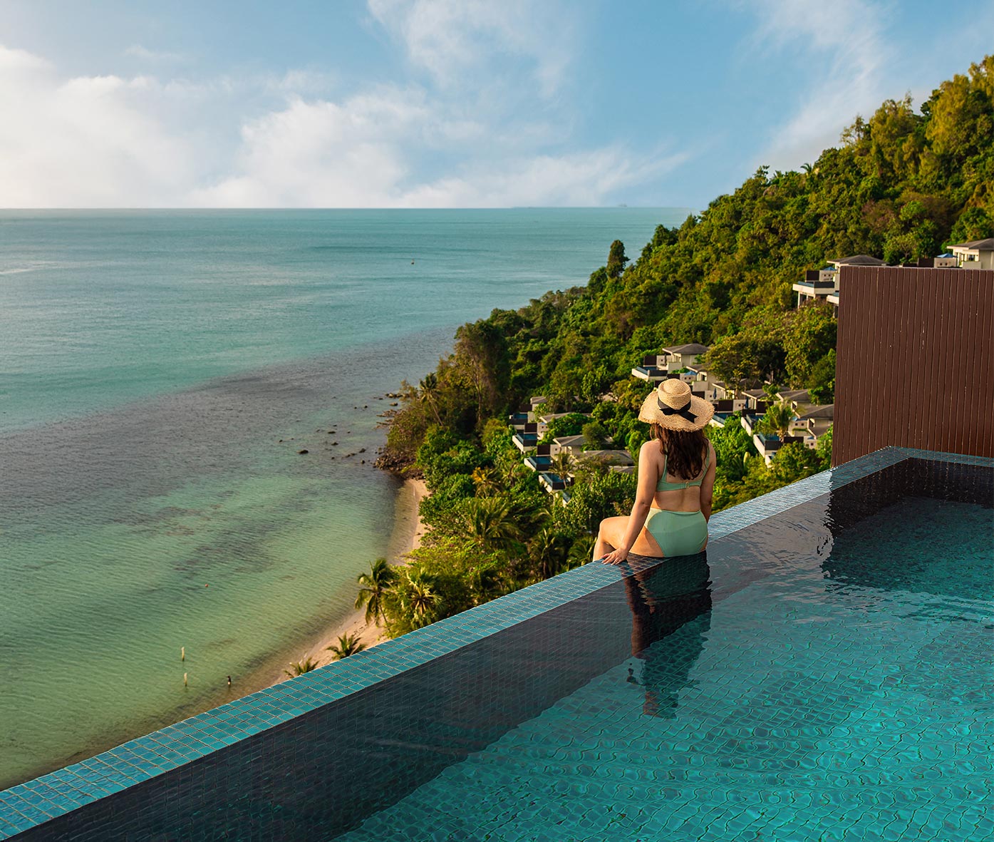 A woman in a light green bikini and straw hat sits at the edge of an infinity pool, overlooking a lush hillside resort and the turquoise ocean stretching to the horizon.







