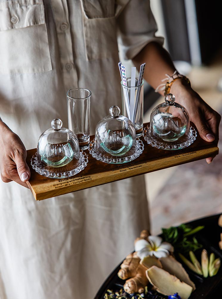 A spa therapist holds a wooden tray with three glass cloches covering essential oils, accompanied by glass vials and scent strips for an aromatherapy treatment. Fresh ginger, tropical flowers, and herbs are arranged on a nearby table, enhancing the luxurious wellness experience.