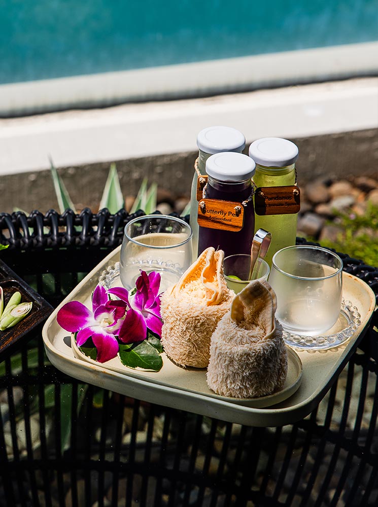 A tray with refreshing welcome drinks, including butterfly pea and pandanus-infused iced tea, is elegantly arranged with two rolled towels, glass cups, and a vibrant purple orchid. The setup is placed on a black wicker table overlooking a tranquil poolside setting.