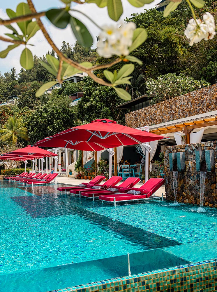 A vibrant outdoor swimming pool features bright red lounge chairs under matching umbrellas, surrounded by lush greenery and a stone waterfall feature. The luxurious poolside setting is complemented by an open-air dining area with white curtains and tropical decor.