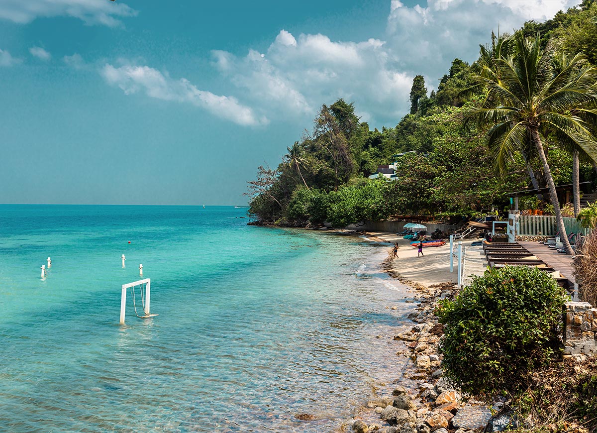 A secluded tropical beach with crystal-clear turquoise waters is lined with lush greenery and swaying palm trees. A partially submerged wooden swing stands in the shallow water, while a few guests relax on the soft sandy shore under beach umbrellas.