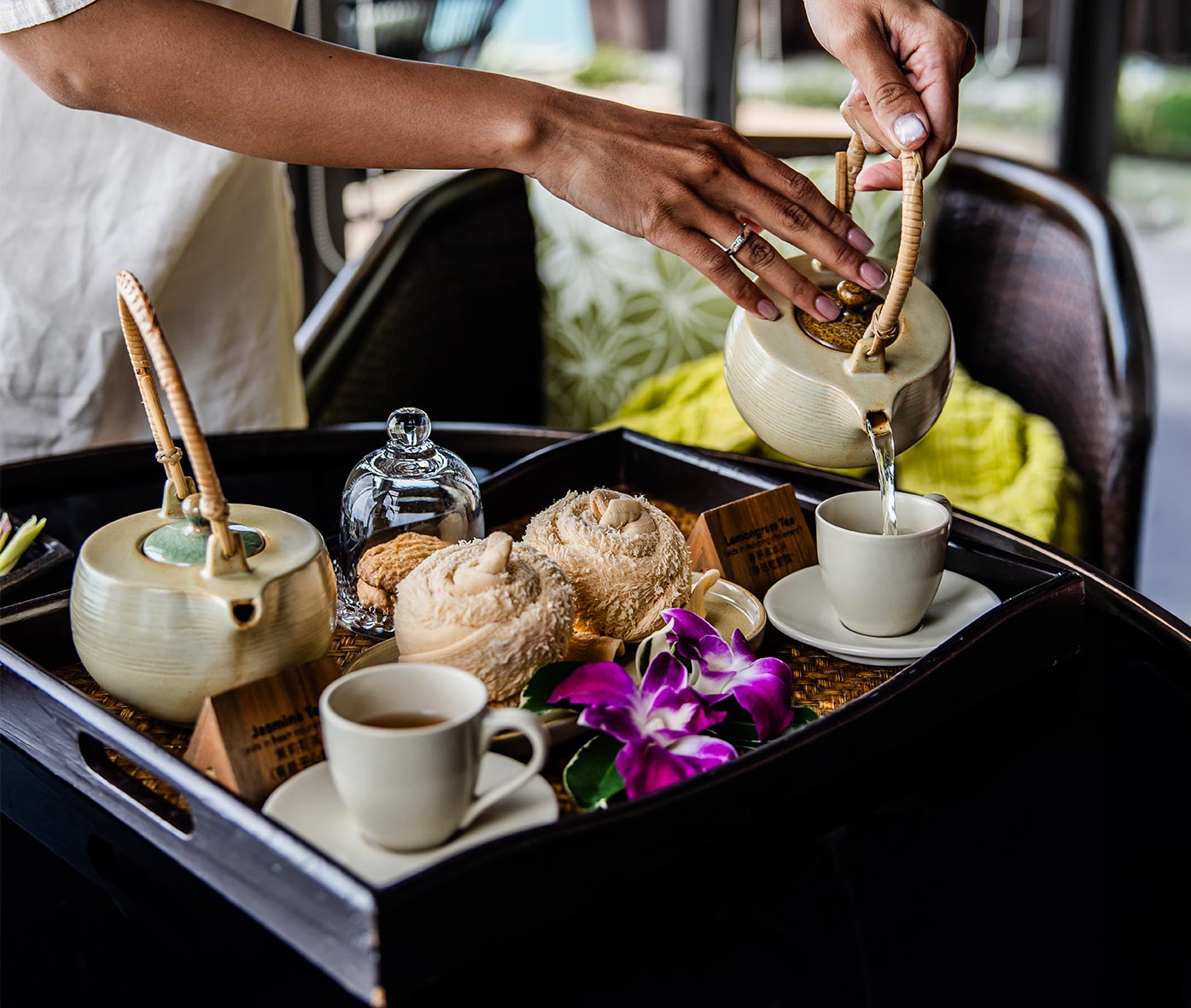 A spa therapist pours tea from a ceramic teapot into a small cup on a beautifully arranged tray featuring rolled towels, a glass cloche with a treat, and a vibrant purple orchid. The elegant setup enhances the serene and luxurious spa experience.