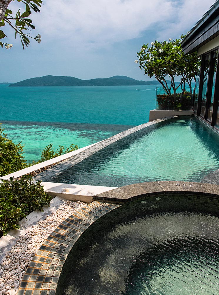 A luxurious infinity pool and circular hot tub overlook the turquoise ocean, with lush greenery and a distant island in the background. The tranquil spa setting blends natural elements with modern design for a serene retreat.