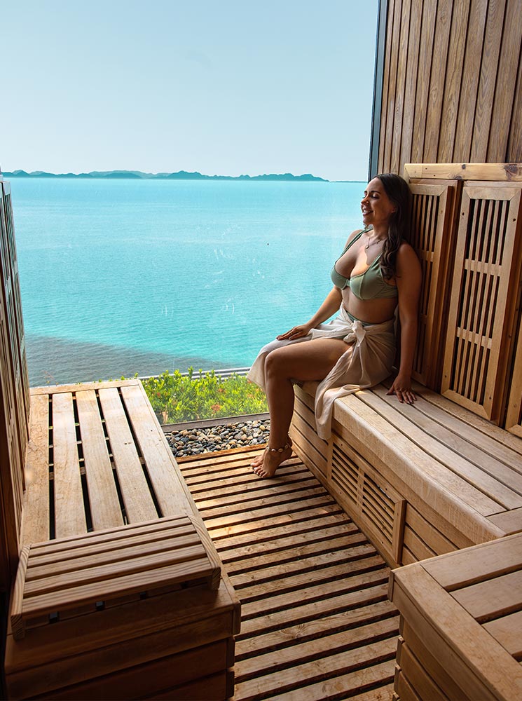 A woman in a green bikini and sheer wrap sits in a wooden sauna, gazing out at the turquoise ocean through a large open window. The natural wood interior and breathtaking sea view create a serene and luxurious spa atmosphere.