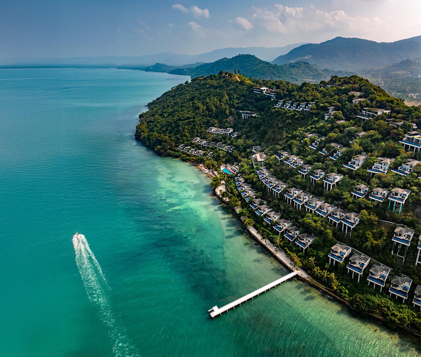 Hotel Review of Conrad Koh Samui a luxury resort in Thailand with private villas on a lush hillside overlooking turquoise waters A white pier extends into the sea and a speedboat leaves a wake behind as it moves through the calm ocean







