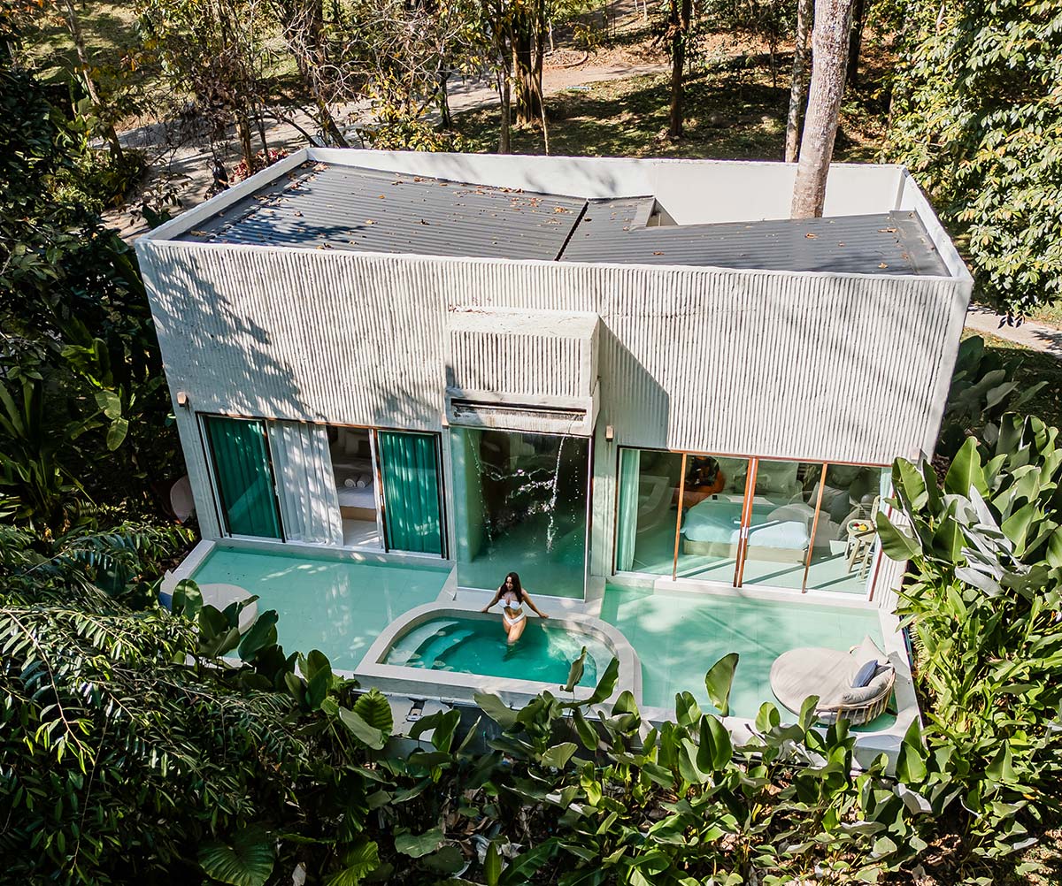 A modern villa with a minimalist design, nestled in lush greenery, featuring a private pool and a cascading water feature. A woman in a white swimsuit relaxes in the pool, enjoying the serene ambiance of the tropical surroundings. Large glass windows and sliding doors connect the sleek indoor space with the tranquil outdoor retreat.







