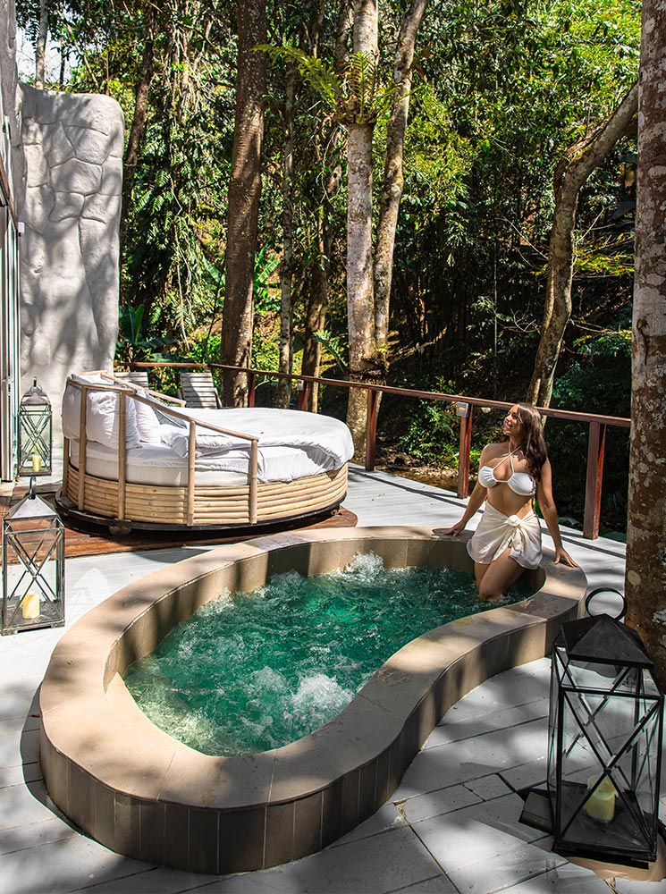 A woman in a white swimsuit sits on the edge of a bubbling jacuzzi on a private outdoor deck, surrounded by lush greenery. A cozy round daybed with plush cushions rests nearby, offering a tranquil spot for relaxation. Soft sunlight filters through the tall trees, enhancing the peaceful, luxurious atmosphere of this secluded retreat.