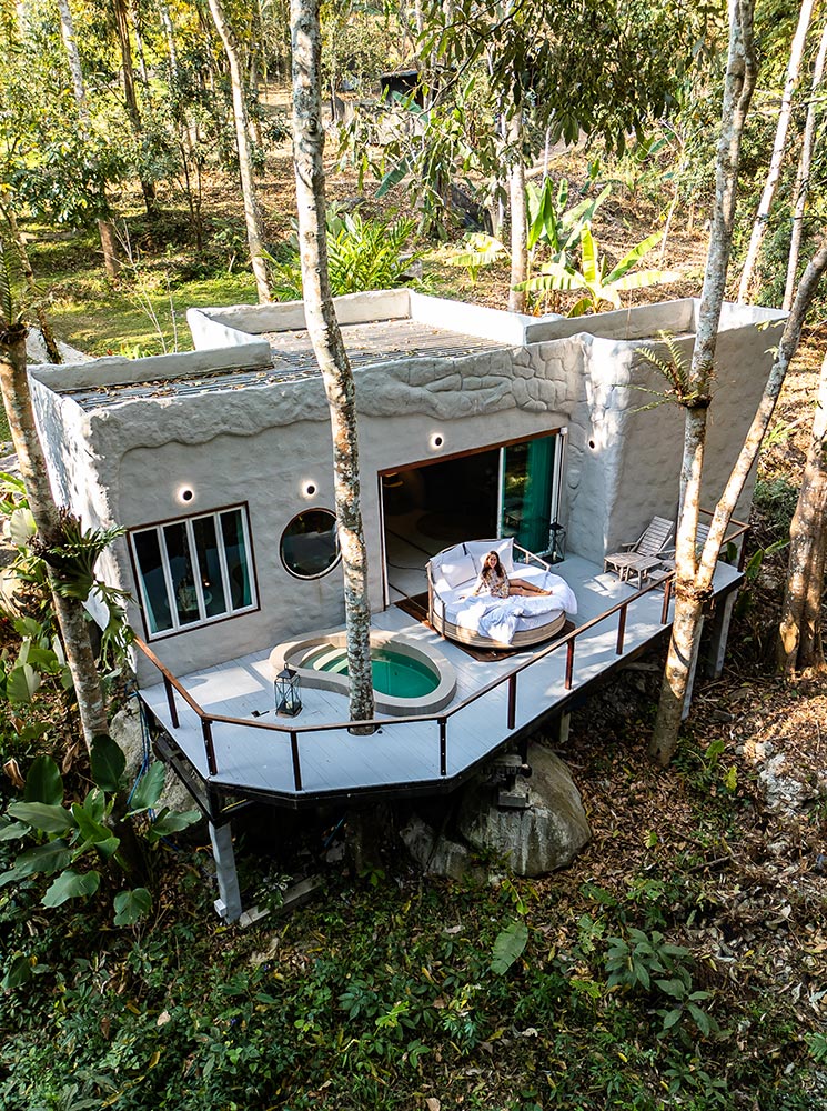 A secluded eco-luxury villa with a unique, textured exterior, nestled among trees in a lush forest. A woman lounges on a round cushioned daybed on the elevated wooden deck, which features a private jacuzzi and modern outdoor seating. The tranquil setting blends seamlessly with nature, offering a peaceful retreat in the heart of Chiang Mai.