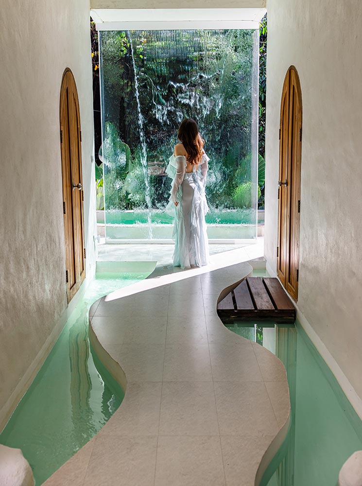 A woman in an elegant white dress stands at the edge of a tranquil, open-air hallway, gazing at a cascading waterfall feature. The curved pathway is lined with shallow water, creating a serene and spa-like atmosphere. Wooden arched doors on either side and soft natural lighting enhance the luxurious, minimalist design of this peaceful retreat.