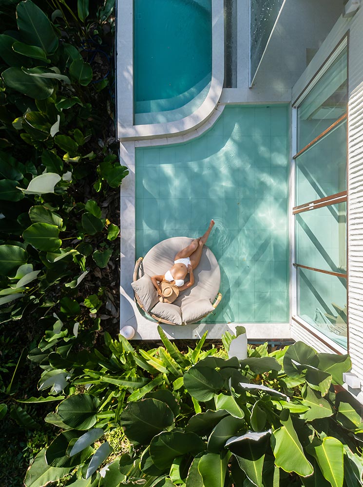 An aerial view of a private poolside retreat surrounded by lush tropical greenery. A woman in a white swimsuit lounges on a round cushioned daybed, holding a sun hat as she relaxes in the sunlight. The modern villa's sleek glass doors open to a turquoise-tiled pool area, creating a seamless blend of indoor and outdoor luxury.