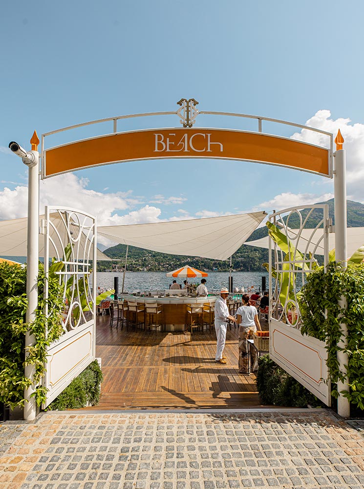 A stylish entrance to a lakeside beach club in Lake Como, Italy, featuring an elegant white and orange archway with the word "BEACH" displayed at the top. Inside, a wooden deck leads to a chic open-air bar with white barstools and a vibrant orange-striped umbrella, overlooking the calm lake waters and surrounding mountains. Two staff members in white attire converse near the entrance, adding to the exclusive and luxurious ambiance.