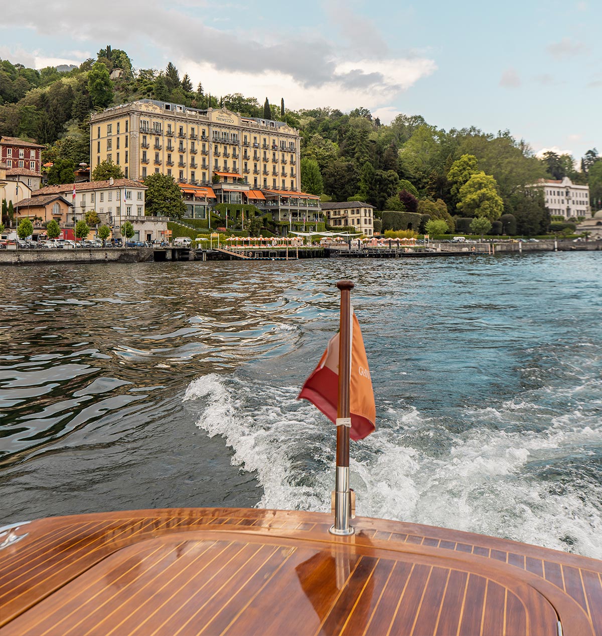 A scenic boat ride on Lake Como, Italy, with a view of the luxurious Grand Hotel Tremezzo along the waterfront. The polished wooden deck of the boat and a fluttering orange flag add to the elegance of the experience. The hotel’s orange umbrellas line the shore, surrounded by lush greenery and historic buildings.