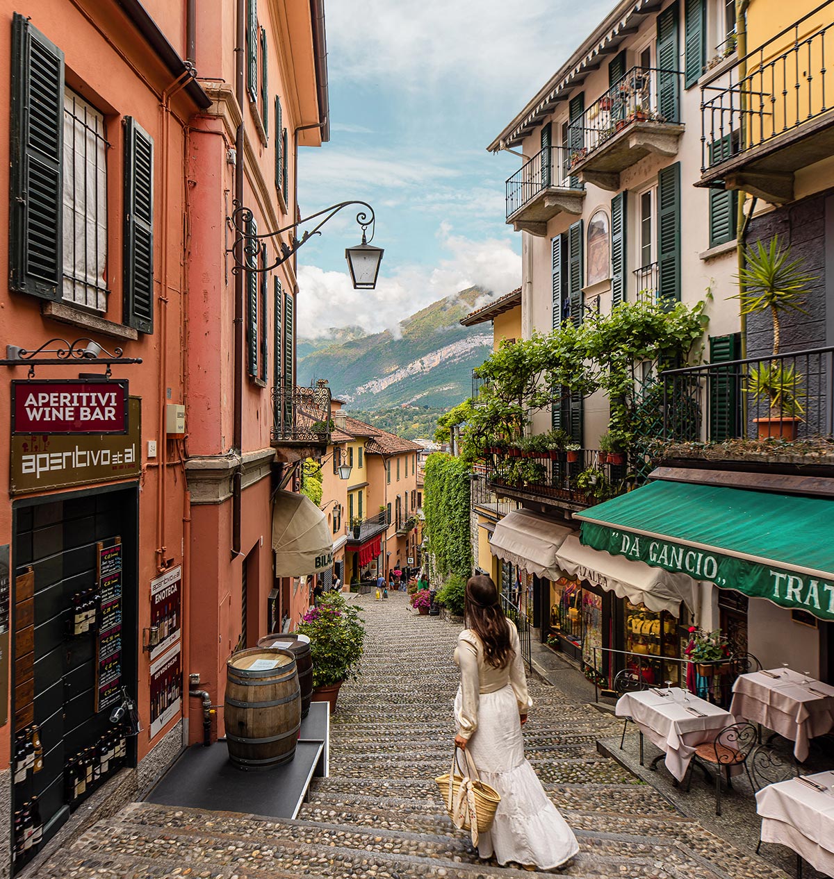 A woman in a flowing white dress walks down a charming cobblestone street in Bellagio, Lake Como, Italy. The picturesque alleyway is lined with colorful buildings, wine bars, and trattorias, with lush greenery and balconies overlooking the descending steps. In the background, the mountains and lake create a breathtaking view.